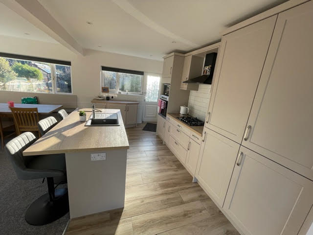 Blossom Avenue Richmond in Matt Cashmere with Natural Halifax Oak Worktop throughout. An island seating three is central with a small row of base units to the left under a window that angle slightly to the door in the back left corner of the room. At the back is a row of units consisting of a tall oven unit, hob with extractor above, integrated fridge freezer and larder unit.