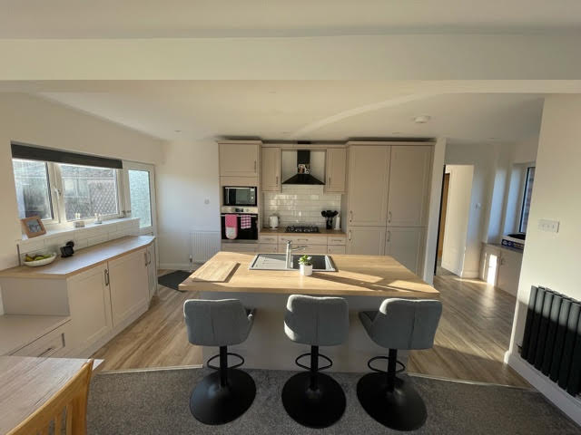 Blossom Avenue Richmond in Matt Cashmere with Natural Halifax Oak Worktop throughout. An island seating three is central with a small row of base units to the left under a window that angle slightly to the door in the back left corner of the room. At the back is a row of units consisting of a tall oven unit, hob with extractor above, integrated fridge freezer and larder unit.