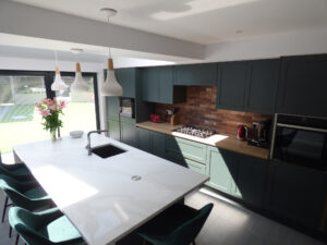 A green handleless Kitchen with brass rails. A wood worktop runs along the back bank with a hob built in. A white quartz worktop sits on the island containing a sink, pop up socket and a large open L-shape seating area.